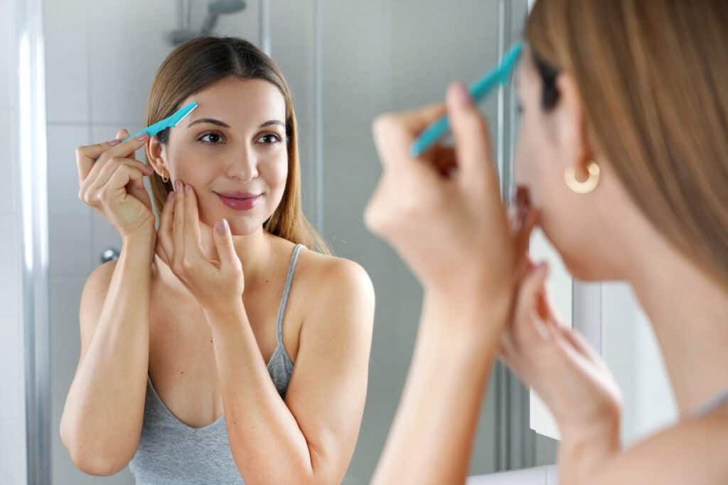 Facial hair removal. Beautiful young woman shaving her face by razor at home. Pretty woman using razor on bathroom. - Beautygab