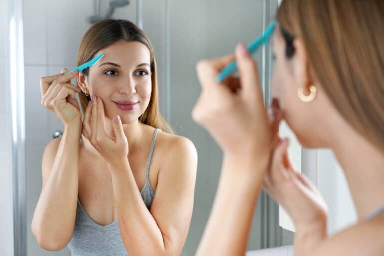 Facial hair removal. Beautiful young woman shaving her face by razor at home. Pretty woman using razor on bathroom.