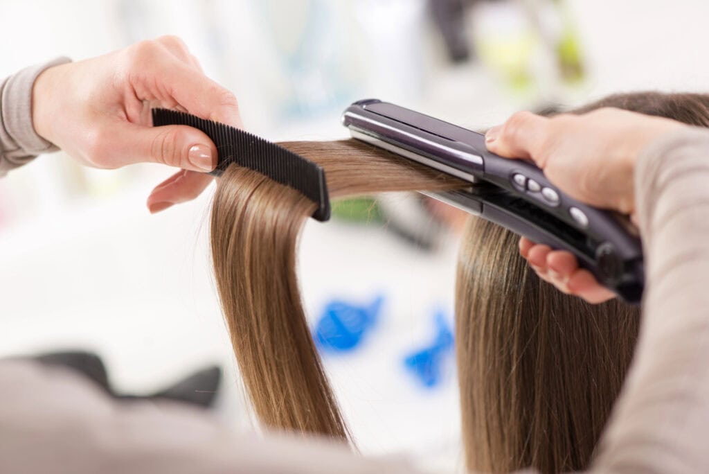 Hair Irons - Beautygab Stylist running a flat iron down a section of a client's long brown hair in a salon