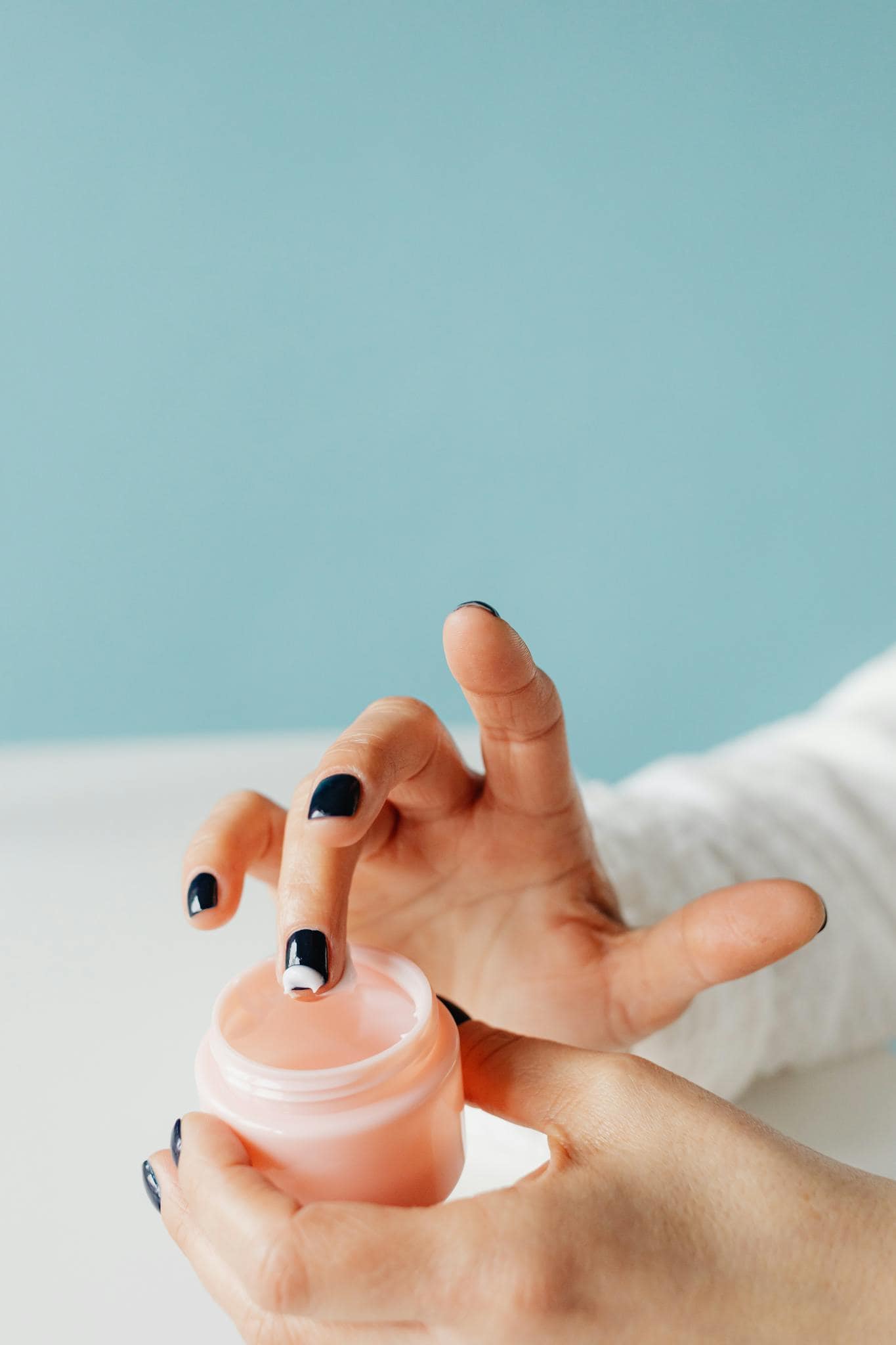 Photo by Karolina Grabowska www.kaboompics.com - Beautygab A close-up of hands applying moisturizing skincare cream from a pink jar against a light blue background.