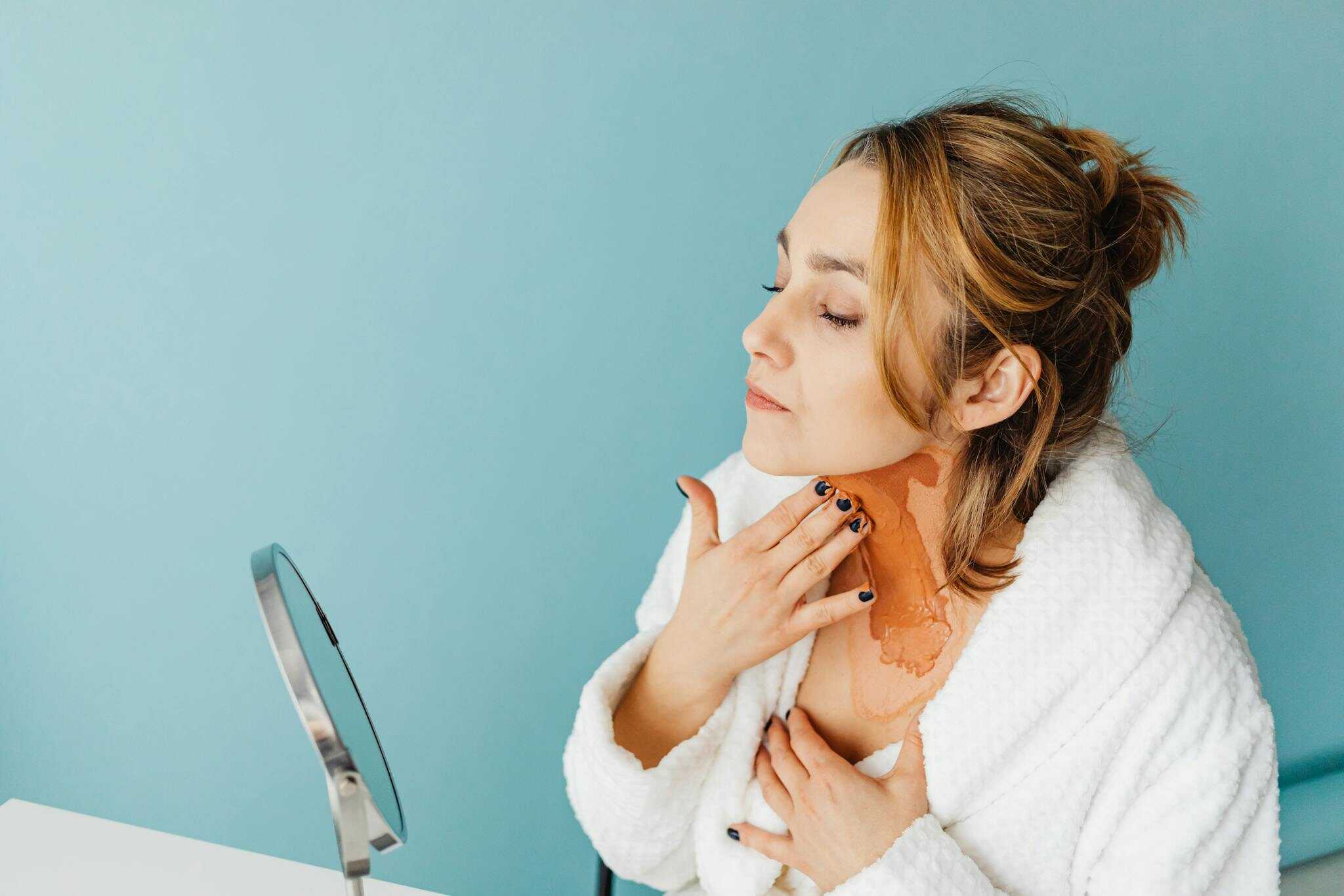 Photo by Karolina Grabowska www.kaboompics.com - Beautygab Woman in a white robe applies skincare product in a studio with a blue background.